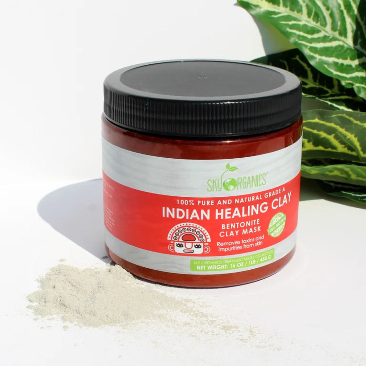 Jar of Indian Healing Clay with a black lid on a white surface with green leaves in the background