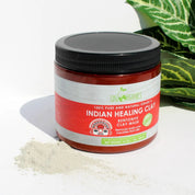 Jar of Indian Healing Clay with a black lid on a white surface with green leaves in the background